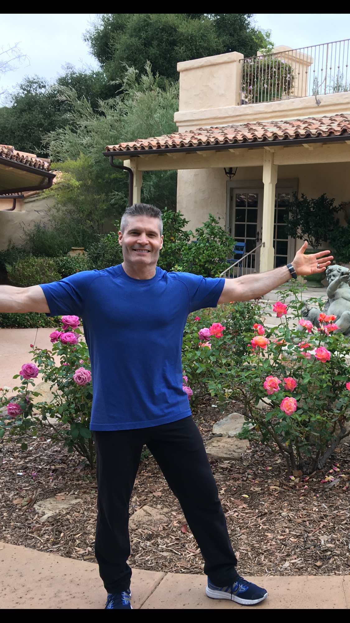 John Turk outside a client’s home, arms open, roses in bloom behind him