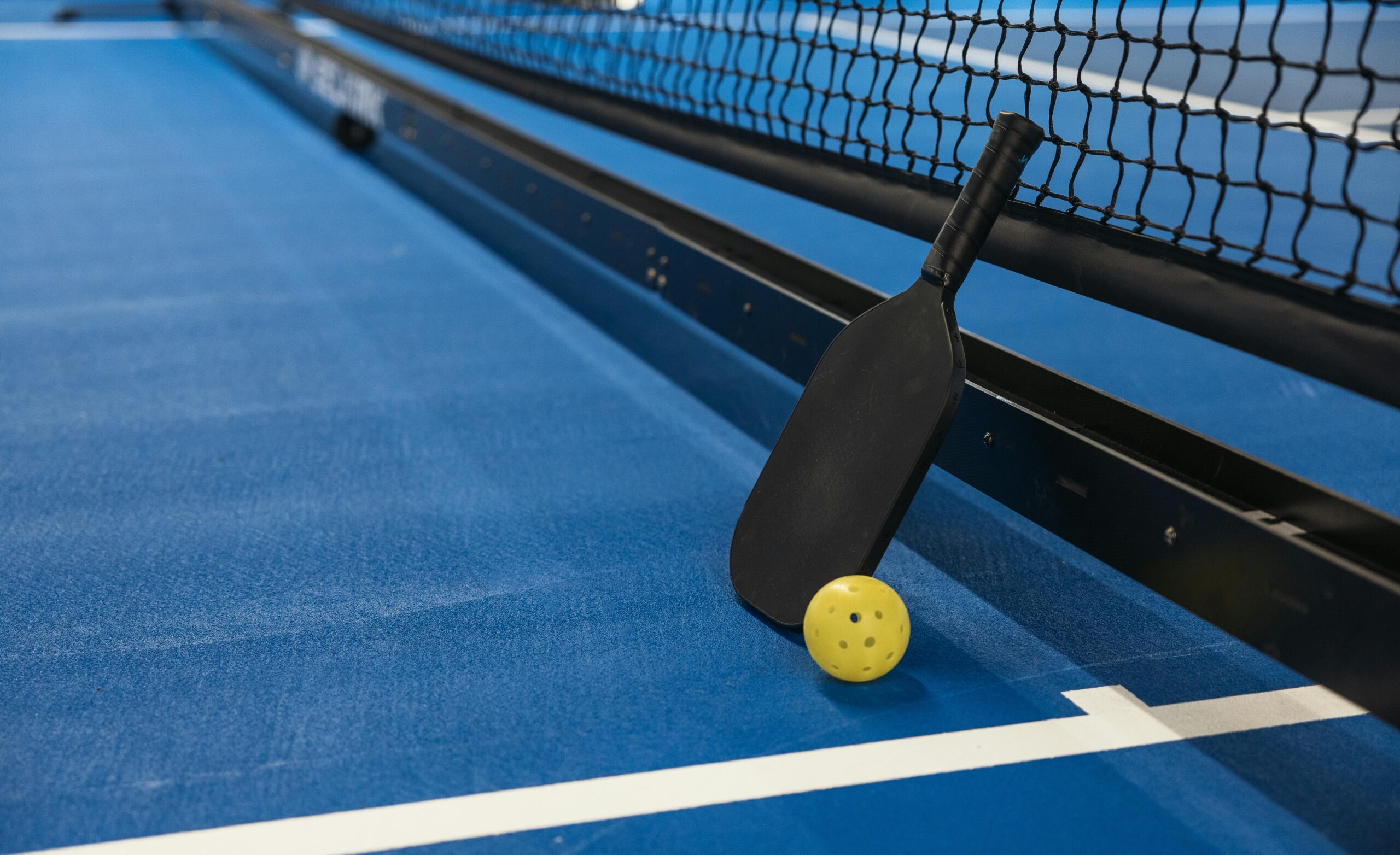 A pickleball paddle and yellow ball resting against the net on a blue court